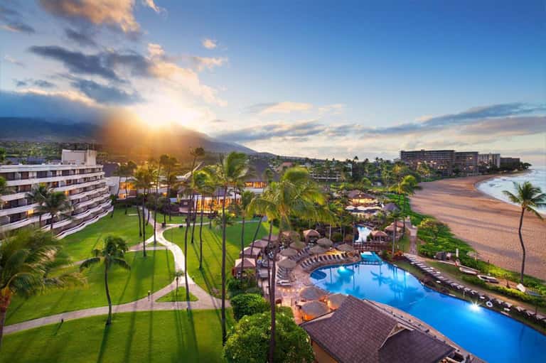 Beachfront resort at golden hour with curved hotel buildings, palm trees, blue pool, and sandy beach