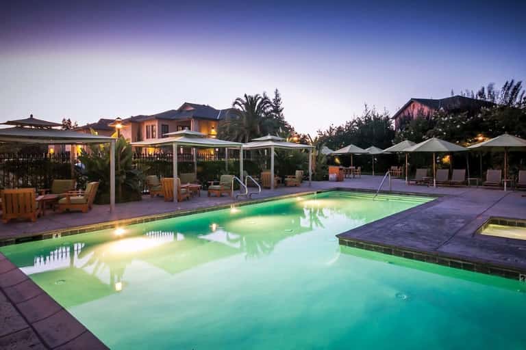 Illuminated resort pool at dusk with white pergolas, lounge chairs, palm trees, and multi-story buildings