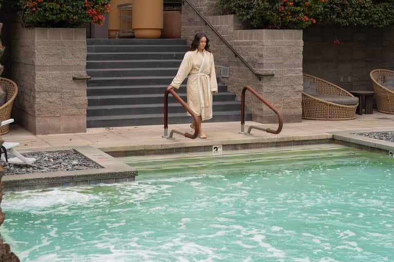 Woman in white robe walking down steps toward a mineral spa pool surrounded by architecture