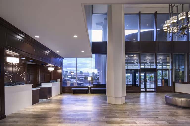 Modern hotel lobby with high ceilings, dark wood reception desk, large windows, polished floors, and contemporary lighting