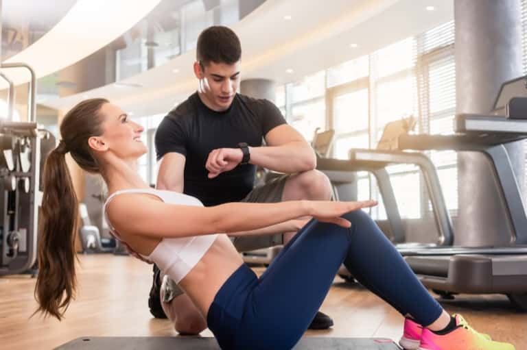 Personal trainer assisting woman doing sit-ups in a bright, modern gym with equipment