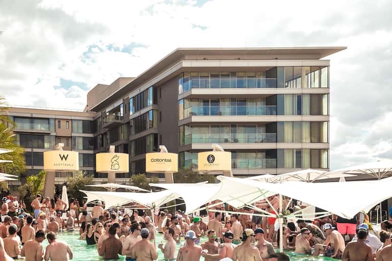 Daytime pool party with crowds swimming, white umbrellas, modern hotel building, and palm trees