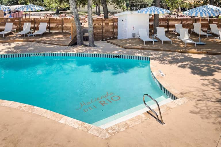 Swimming pool with 'Hacienda Del Rio' text, white lounge chairs, blue umbrellas, and wooden fence