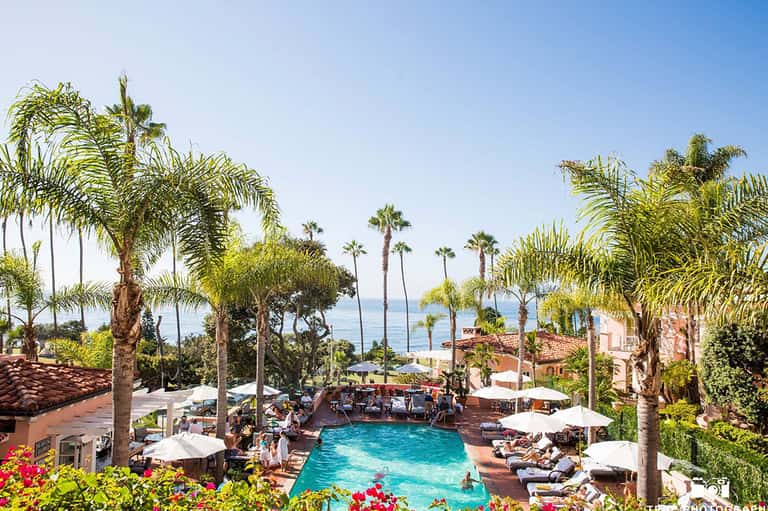 Aerial view of resort pool surrounded by palm trees, white umbrellas, lounge chairs, and ocean view in background