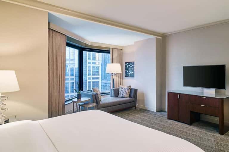 Modern hotel bedroom with king bed, sofa seating area, and floor-to-ceiling windows