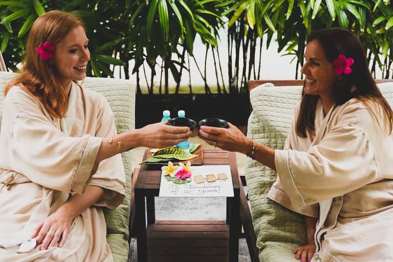 Two women clinking glasses at a spa, wearing robes with tropical plants in background