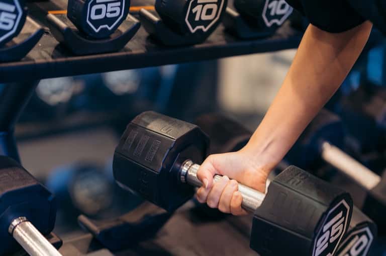 Close-up of woman's hand holding black dumbbell in fitness gym with weight rack background