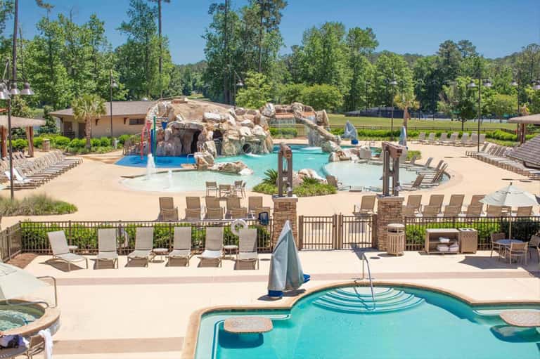 Expansive resort pool area with water feature, rock formation, multiple pools, lounge chairs, and wooded surroundings