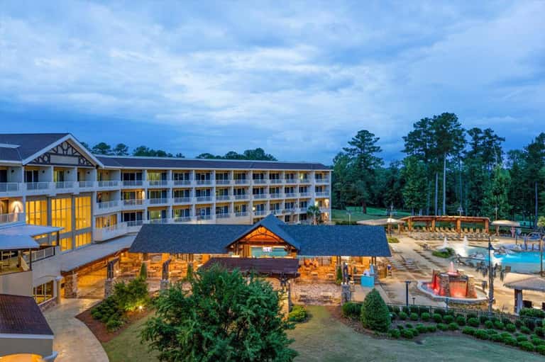Luxury resort exterior at dusk with illuminated main lodge, white columns, multiple balconies, and lit pool area.