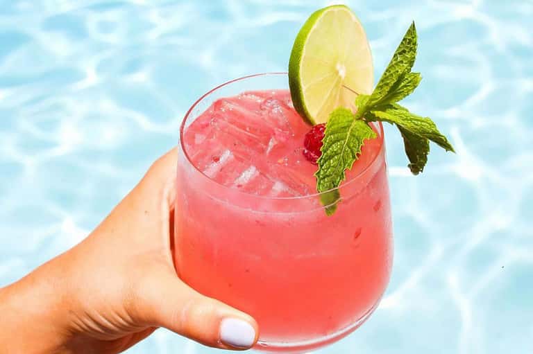 Tropical cocktail with coral-colored liquid, lime slice, and mint garnish held over pool water