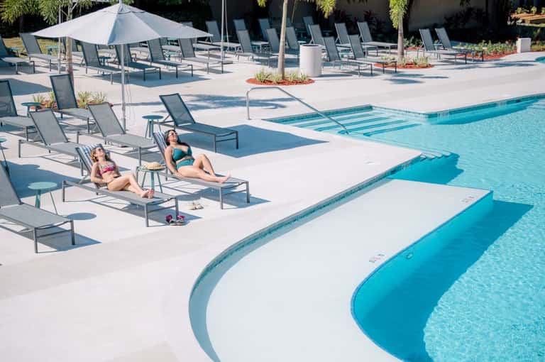 Resort pool area with guests lounging on chairs, white umbrellas, and clear blue water