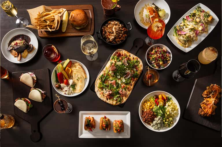 Resort dining spread with tacos, burgers, seafood dishes, cocktails, and appetizers on black table