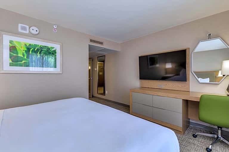 Hotel room with queen bed, TV mounted above wooden dresser, green chair, and digital art display