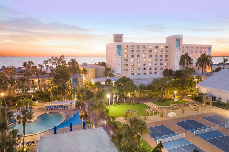 Beachfront resort at sunset with hotel towers, pool, tennis courts, and palm trees