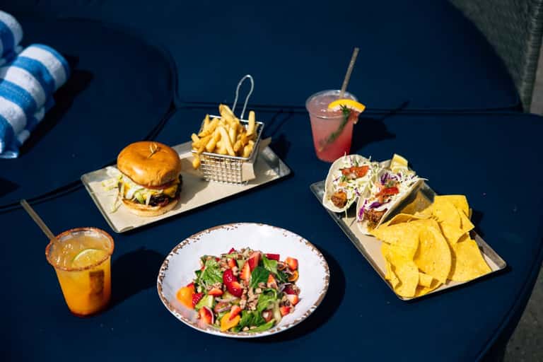 Poolside food and drinks including burger, fries, tacos, salad, and colorful cocktails on blue table
