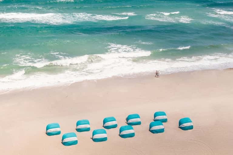 Aerial view of beach umbrellas on sand with turquoise ocean and surfers in the water