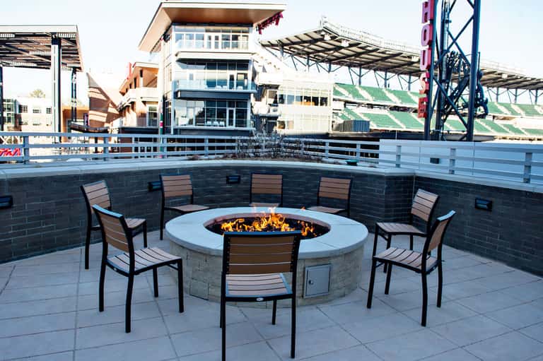 Modern outdoor fire pit lounge area with seating surrounded by sports stadium and buildings