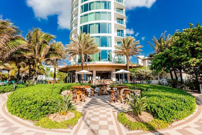 Resort exterior with curved white building, tropical landscaping, palm trees, and wooden seating area