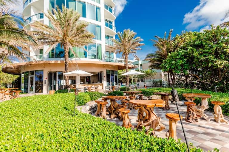Beachfront resort grounds with wooden seating areas, palm trees, lush lawn, and building with umbrellas