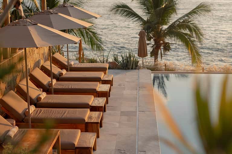 Beachfront lounge area with cushioned sunbeds under umbrellas, palm trees, and ocean view at golden hour