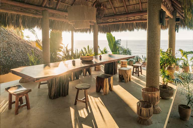 Beachfront dining pavilion with thatched roof, wooden table, and ocean view at golden hour