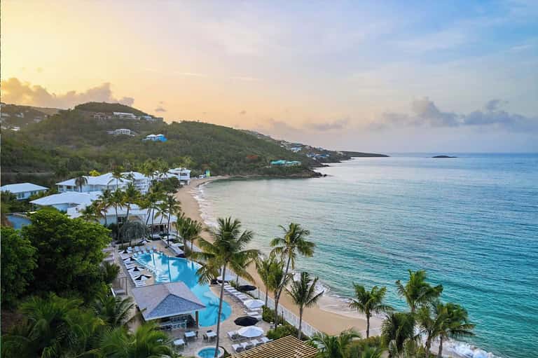Beachfront resort at sunset with pool, palm trees, white buildings on tropical hillside overlooking ocean