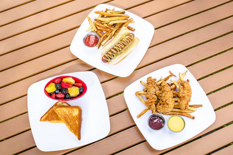 Three white plates with gourmet food including hot dog with toppings, fried chicken with dipping sauces, grilled cheese with fruit, and french frie...