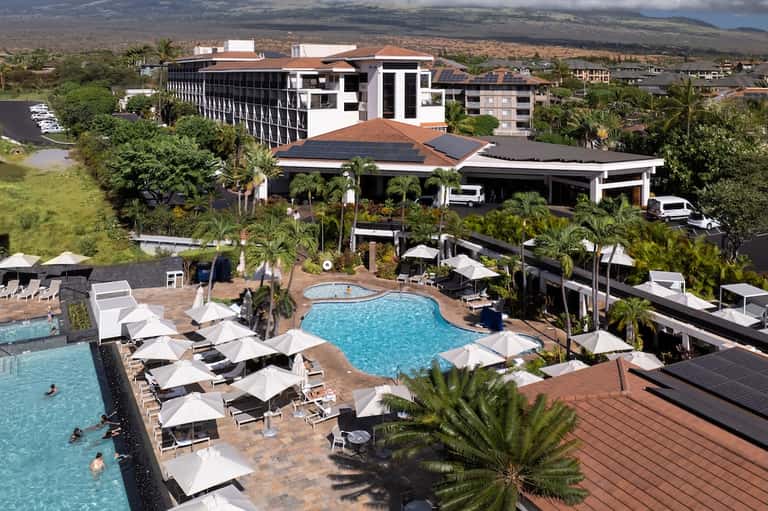 Aerial view of resort with curved pools, palm trees, and white umbrellas overlooking landscape
