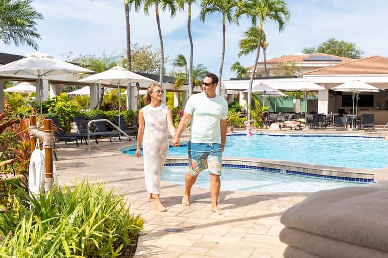 Couple walking along resort pool deck with palm trees, umbrellas, loungers, and tropical landscaping