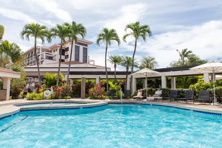Resort pool surrounded by palm trees, white pergolas, lounge chairs, and multi-story residential building