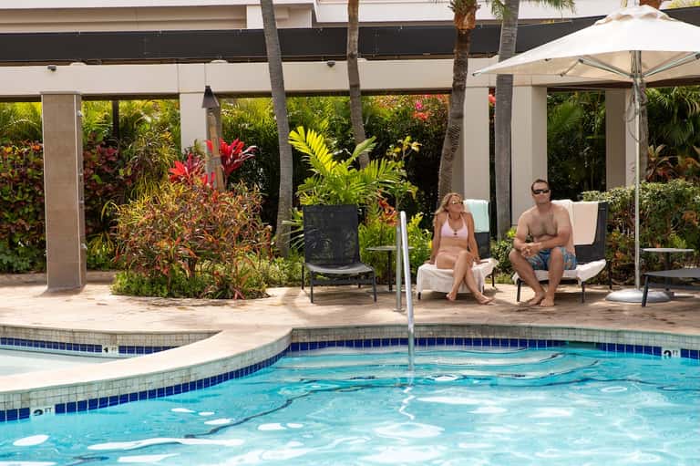 Resort pool with tropical plants, lounge seating under pergola, and guests relaxing poolside