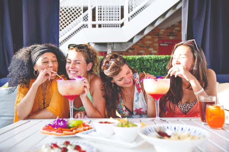 Four women enjoying colorful cocktails and appetizers at an outdoor poolside gathering