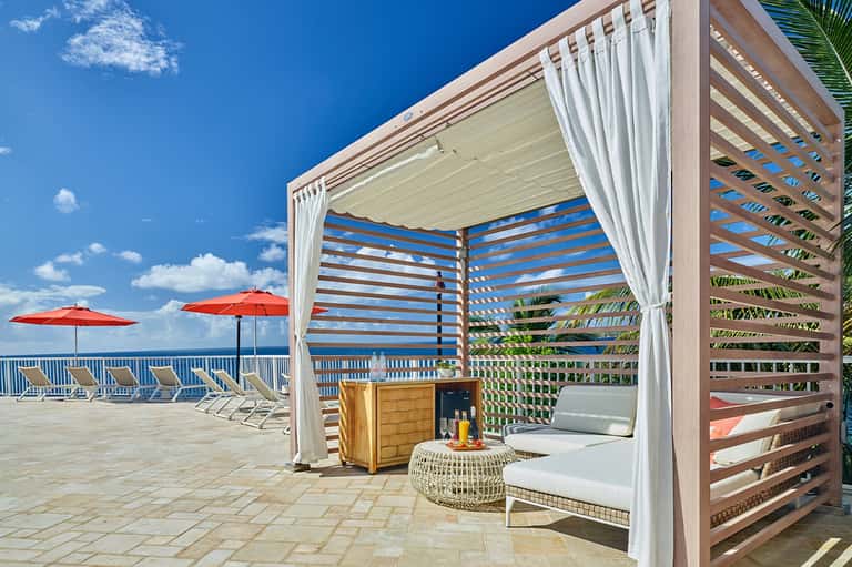 Oceanfront cabana with retractable roof, white curtains, red umbrellas, and lounge seating