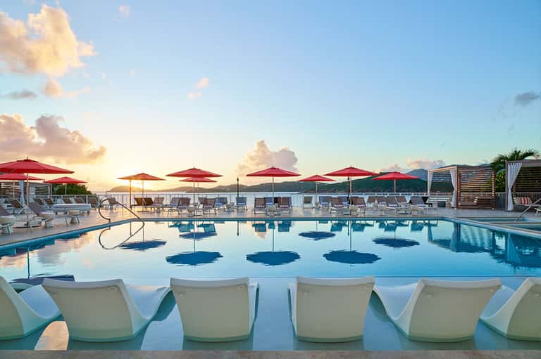Sunset pool scene with red umbrellas, white lounge chairs, and mountains in background