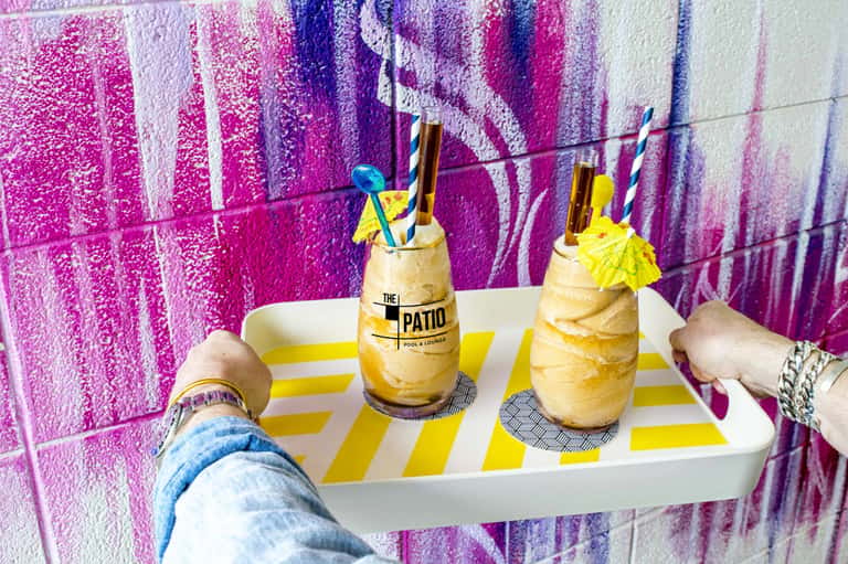 Two tropical cocktails with yellow umbrellas on striped tray against colorful pink and purple textured wall