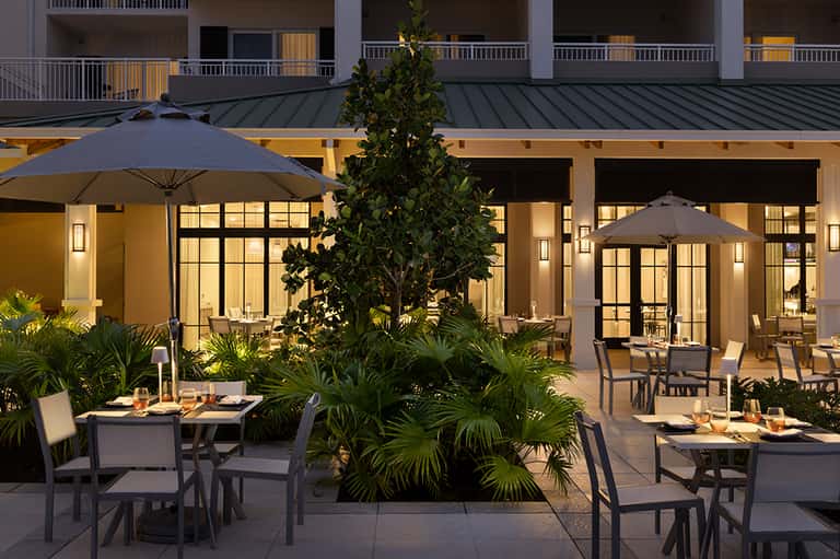 Outdoor dining patio at dusk with dark umbrellas, tropical plants, and warm lighting