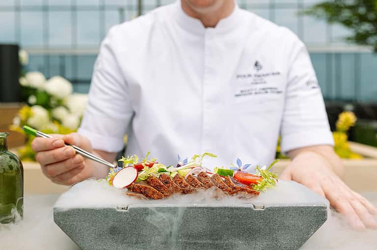 Chef in white jacket presenting plated dish with microgreens and garnish outdoors