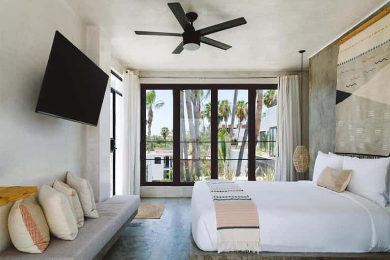 Modern resort bedroom with bed, seating area, flat-screen TV, and sliding glass doors opening to palm tree views