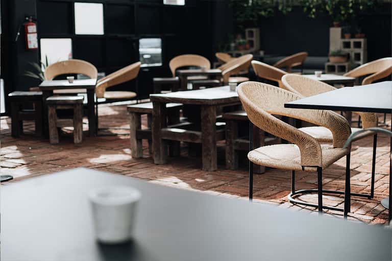 Outdoor terrace dining area with woven natural chairs, white tables, and black glass wall backdrop in afternoon sunlight