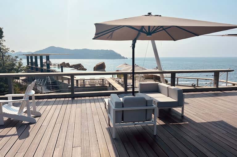 Coastal deck overlooking ocean with white lounge seating, large umbrella, wooden flooring, and island views