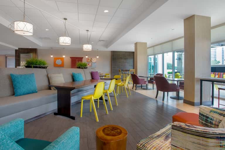 Contemporary hotel lobby with yellow chairs, gray seating, colorful accents, pendant lights, and large windows