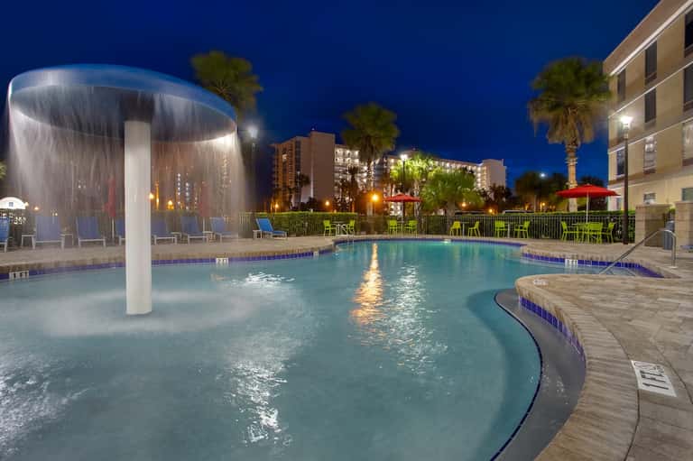 Illuminated resort pool at night with water feature, palm trees, and hotel buildings