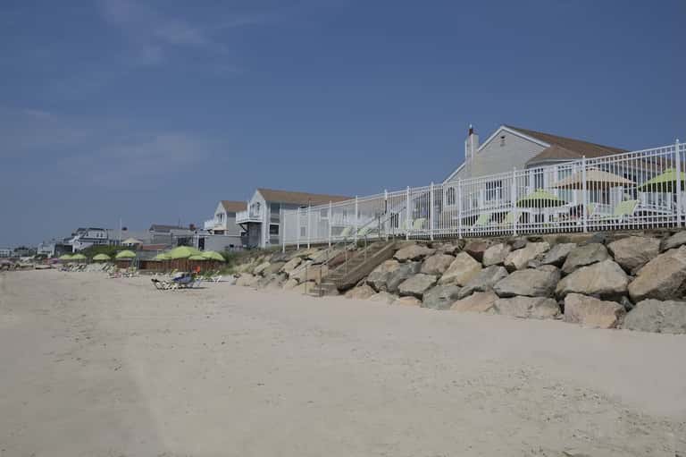 Beachfront homes with white railings on sandy shore protected by large rock breakwater
