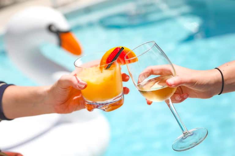 Two hands clinking colorful cocktails by a resort pool with a floatie in the background