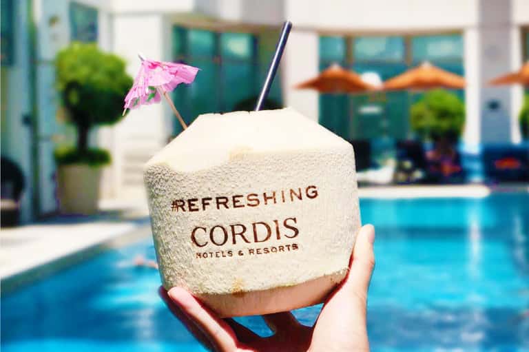Coconut drink with pink umbrella and straw by resort pool with lounge chairs and umbrellas