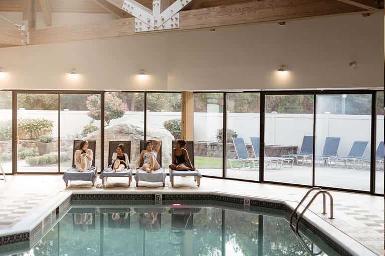 Indoor pool area with lounge chairs, large windows overlooking landscape, and exposed wooden beams