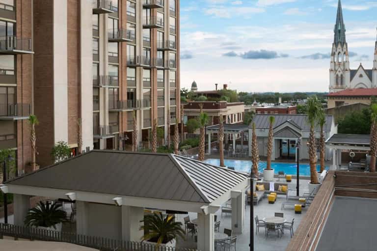Elevated view of resort pool deck with white pergola, palm trees, surrounding hotel buildings, and distant church spire