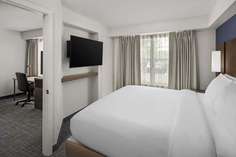 Hotel guest room with white bedding, wall-mounted TV, work desk, chair, and large windows with neutral curtains