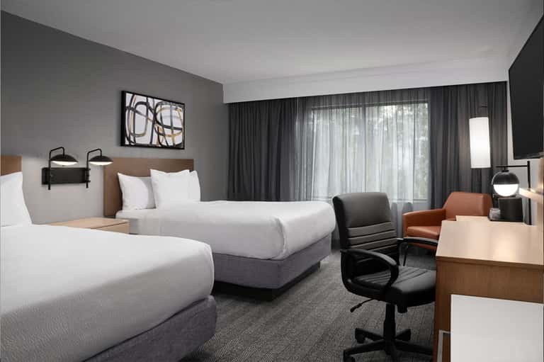 Modern hotel room with two queen beds, work desk, sleek furnishings, and floor-to-ceiling windows.