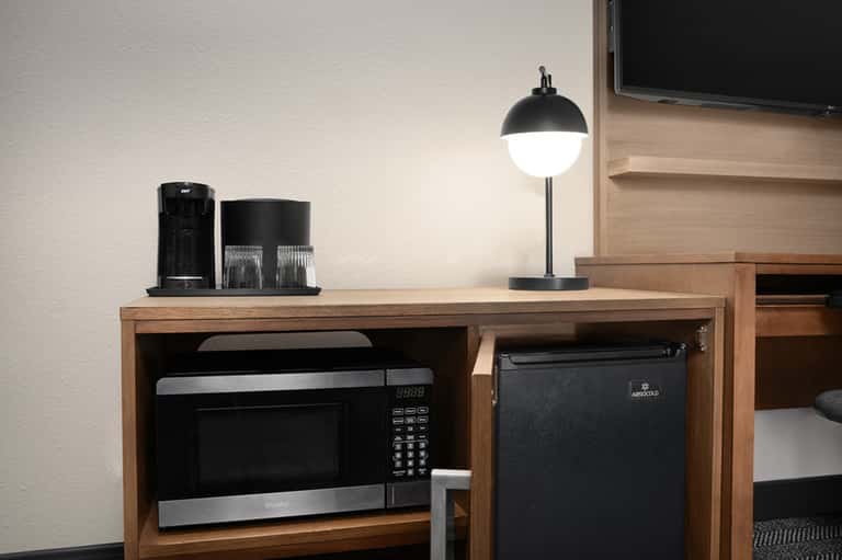 Hotel room desk with coffee maker, mini-fridge, desk lamp, and TV mounted on wooden wall unit